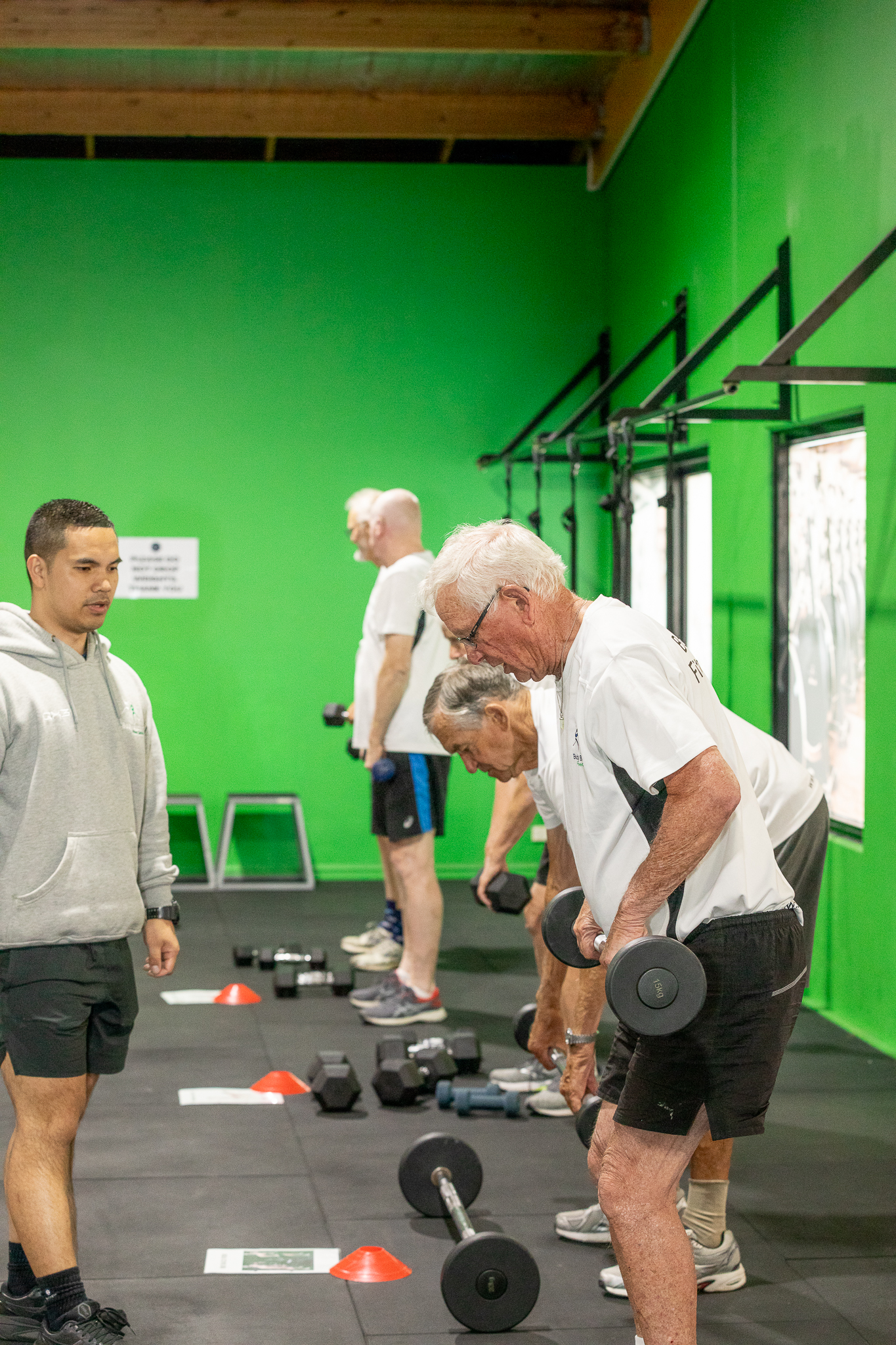 Group of older men exercising with dumbbells in a gym with bright green walls, guided by a young male instructor.